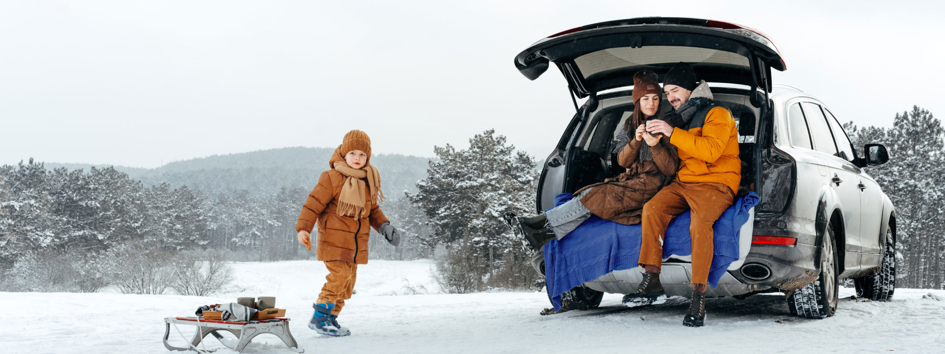Семья, тепло одетая для зимы, сидит в открытом багажнике автомобиля, окруженном заснеженными деревьями. Ребенок, тянущий сани, подходит, улыбаясь.
