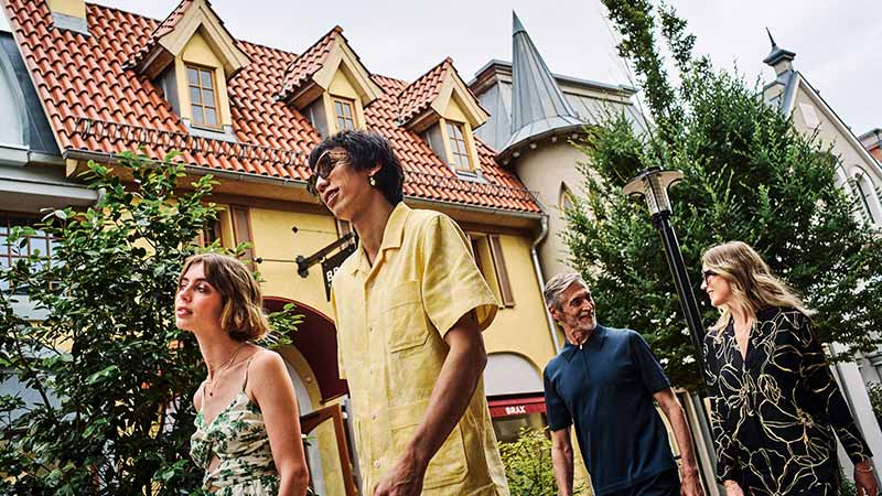 Four people walk outside in front of buildings with steep, red-tiled roofs and dormer windows on a cloudy day.