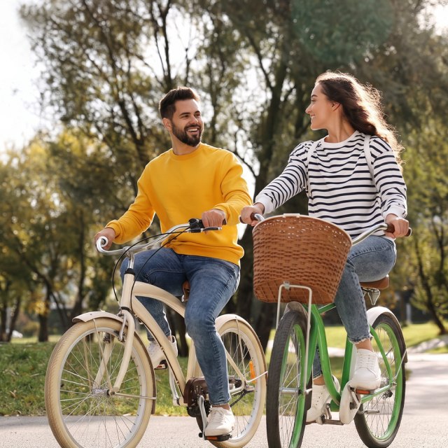 A smiling couple rides bicycles in a sunny park, surrounded by lush green trees.