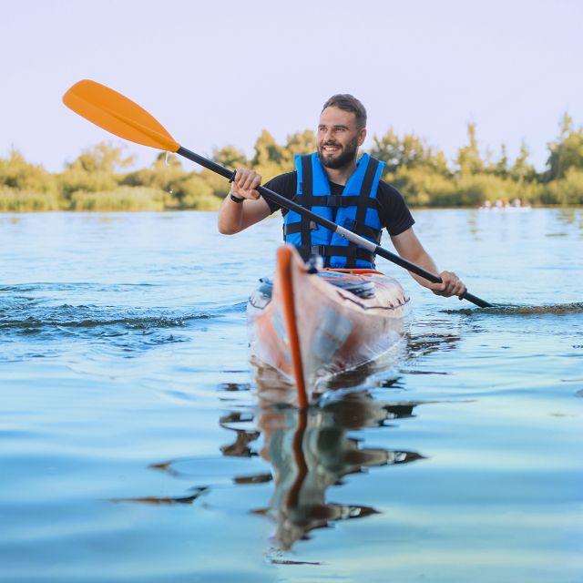 A man in a blue life vest kayaks on a calm lake, holding an orange paddle.