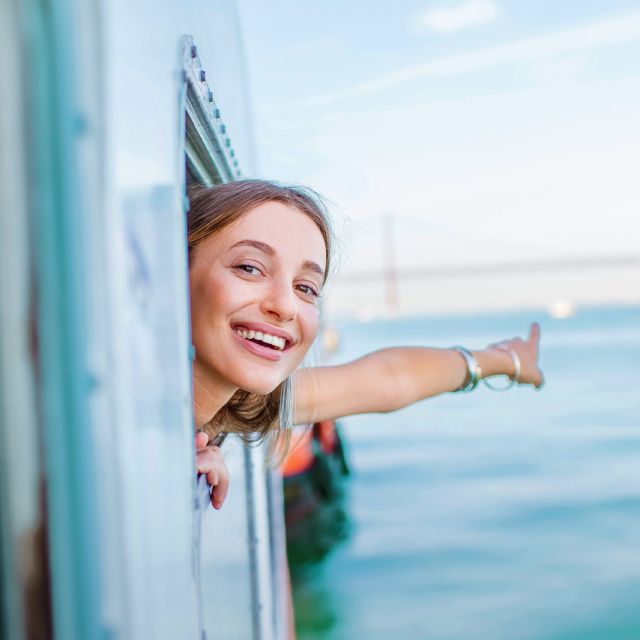 A smiling woman leans out of a train window, joyfully pointing towards the ocean under a clear blue sky.