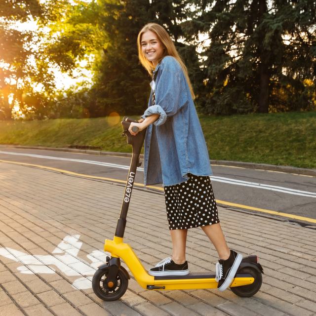 Smiling woman riding a yellow electric scooter on a paved path in a park, wearing a denim jacket and polka-dot skirt.