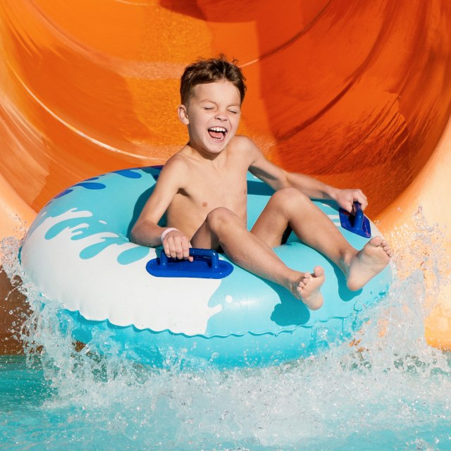 A joyful child slides down an orange water slide in a blue and white tube, splashing into a pool, radiating excitement and fun at a water park.