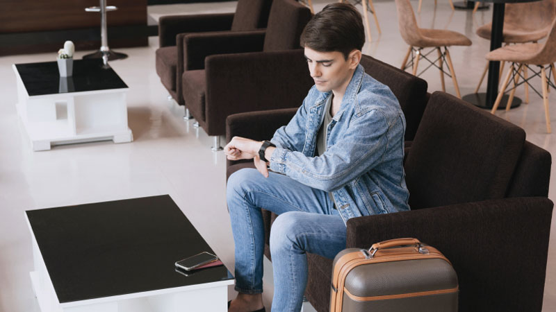 Человек в джинсовой куртке сидит в зале ожидания аэропорта, поглядывая на свои наручные часы. Он выглядит задумчивым. Рядом на столе лежат чемодан и смартфон.