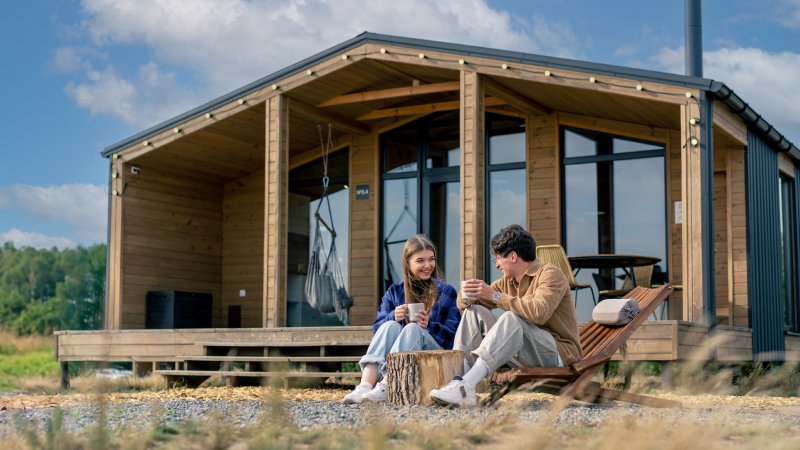 A smiling couple sits outside a modern wooden cabin on a sunny day, enjoying coffee.