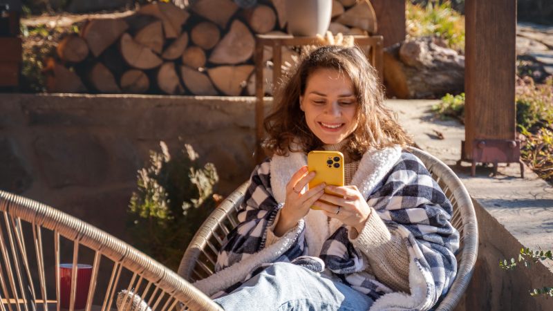 Женщина в уютном клетчатом пледе сидит на улице на плетеном стуле и улыбается своему желтому телефону.