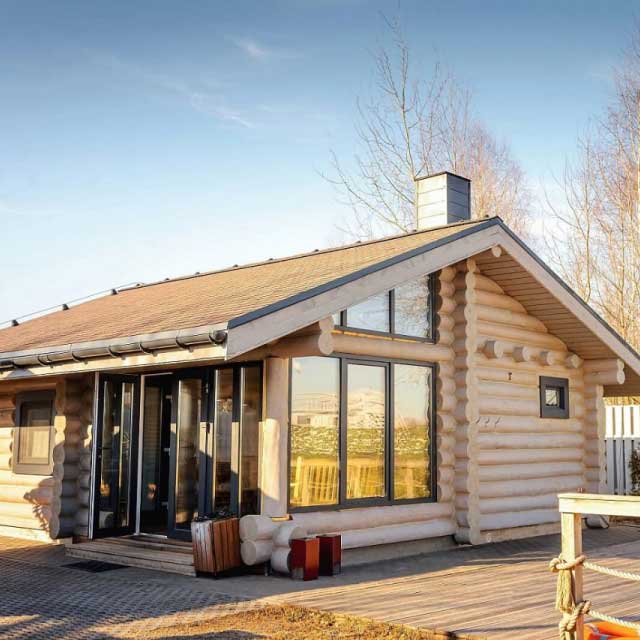 Уютная бежевая бревенчатая хижина с большими окнами и дымоходом стоит под чистым голубым небом.