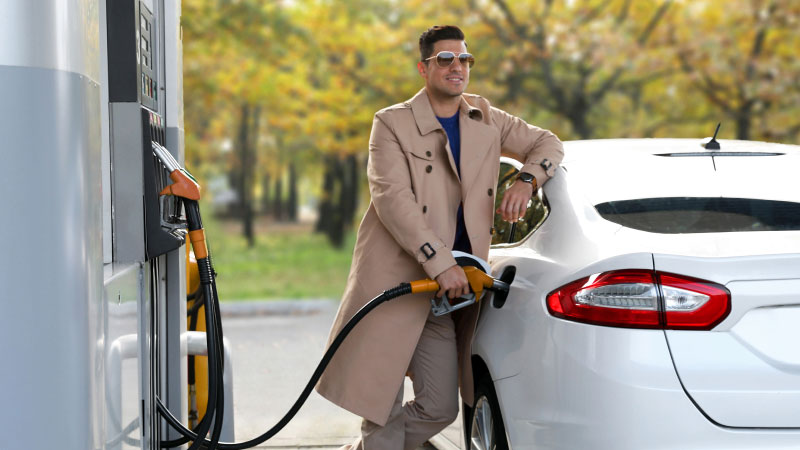 Мужчина в светло-коричневом пальто и солнцезащитных очках улыбается, заправляя белый автомобиль на заправочной станции. На заднем плане видны деревья с осенней листвой.