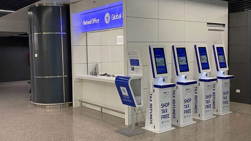 Зона киосков по возврату налога с четырьмя бело-голубыми автоматами с надписью «Shop Tax Free» рядом с стойкой «Refund Office» в аэропорту.