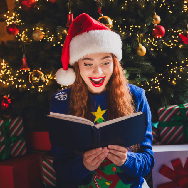 Женщина в шапке Санты и праздничном свитере радостно читает книгу перед украшенной елкой, окруженный завернутыми подарками.