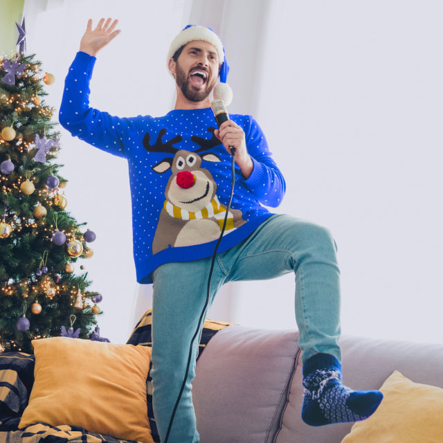 Мужчина в праздничном свитере с оленями и шапке Санты поет в микрофон.