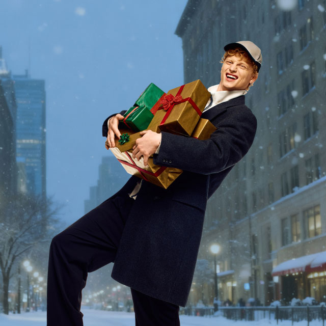 Веселый человек в пальто и шапке несет красочные, завернутые подарки по заснеженной улице города