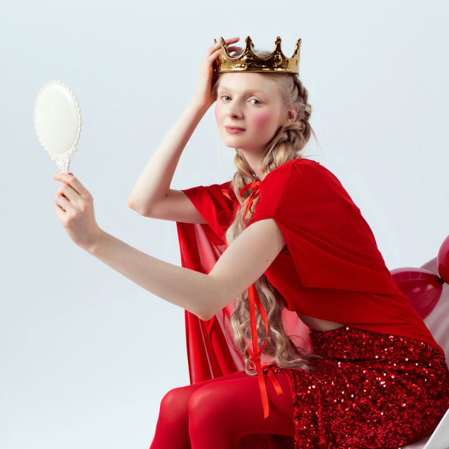 Женщина в красном платье сидит с изяществом, на голове у нее золотая корона, в руках она держит богато украшенное зеркало. 