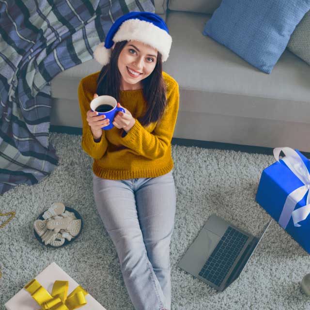 Женщина в синей шапке Санты и желтом свитере сидит на ковре, держа в руках кружку. Рядом с ней лежат ноутбук, подарки и печенье.