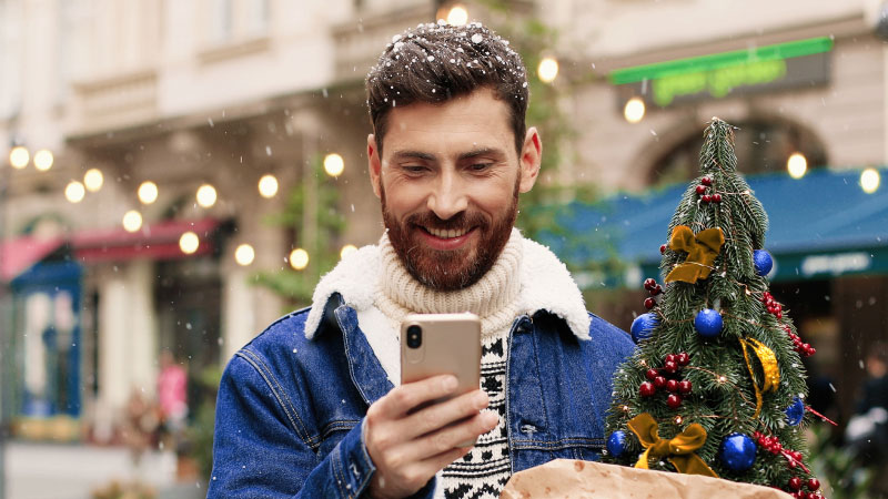 Бородатый мужчина в уютном свитере и джинсовой куртке улыбается, глядя на свой телефон. Он держит в руках небольшую украшенную елку.