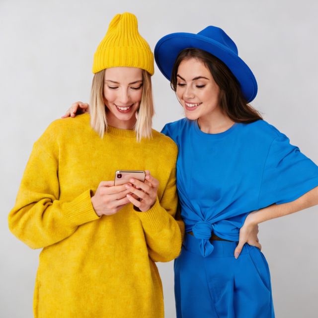Две улыбающиеся женщины смотрят на телефон. Одна в желтом свитере и шляпе, другая в синем платье и шляпе.