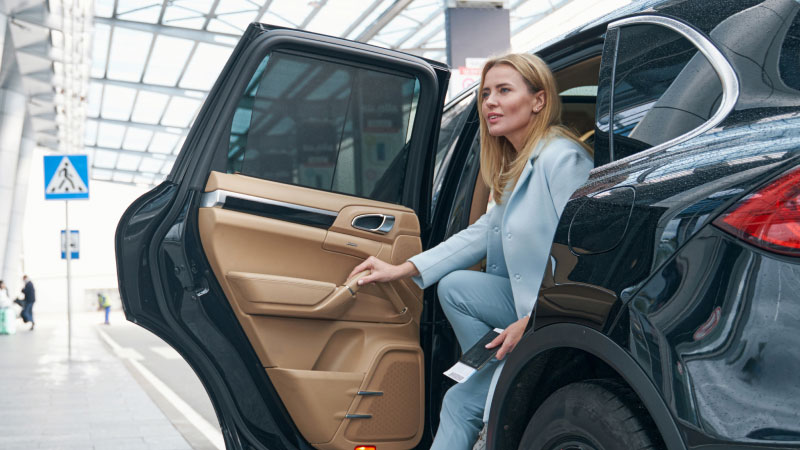 Женщина в голубом костюме выходит из черного автомобиля в терминале аэропорта. 