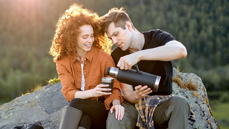 Пара сидит на камне на природе, окруженная природой, улыбается. Мужчина наливает напиток из термоса. Солнечный свет проникает сквозь деревья, создавая теплую, умиротворенную атмосферу.