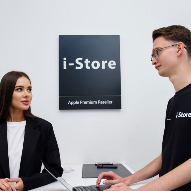 Двое людей стоят у прилавка в магазине Apple Premium Reseller с ноутбуком. Мужчина одет в футболку с надписью «i-Store», женщина - в блейзер.