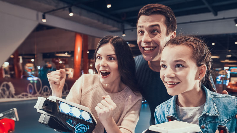A group of three people, including two young women and a man, play arcade games together and appear excited.