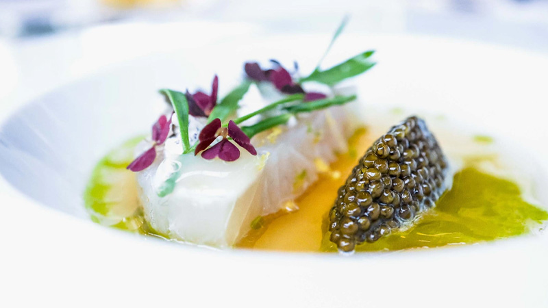 A white fish fillet garnished with microgreens and edible flowers, served with a spoonful of caviar and sauce in a white dish.