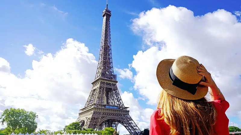 A woman in a straw hat and red shirt looks up at the Eiffel Tower on a sunny day with blue sky and scattered clouds.