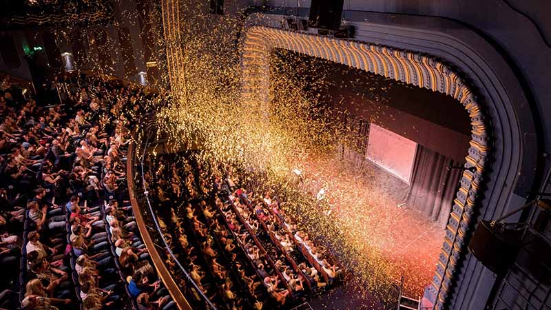 A theater audience watches a stage performance as gold confetti falls from above, filling the air over the crowd and stage.