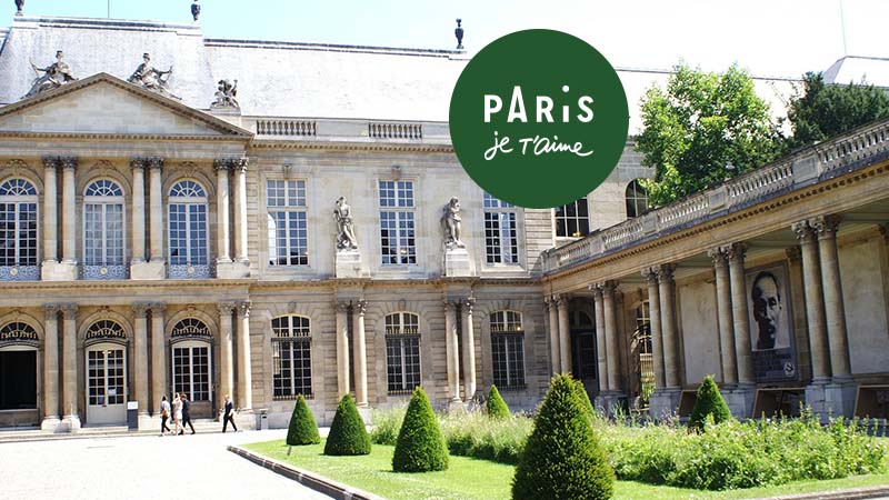 Courtyard of a historic stone building with arched windows, manicured bushes, and a green circular "Paris je t’aime" logo at the top center.