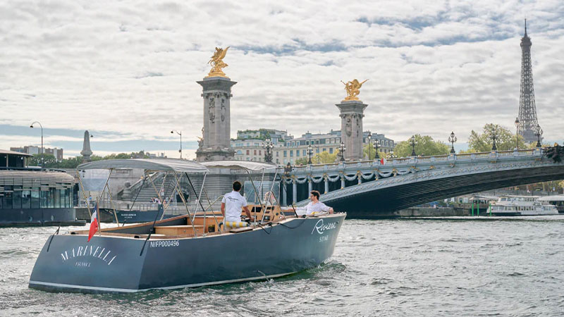 A motorboat with two people sails on the Seine River in Paris, passing by the Pont Alexandre III bridge with the Eiffel Tower visible in the background.