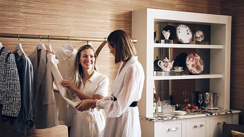 Two women stand in a room with a clothing rack, looking at clothes and smiling. Shelves with dishes, glassware, and fruit are visible in the background.