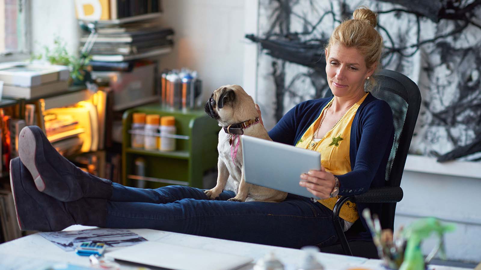 Une femme avec ses pieds posés sur un bureau, lisant une tablette, un chien sur ses genoux.